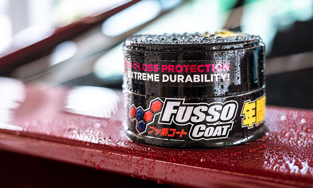 Photo of a Soft99 car care product Fusso Coat 12 Months wax, sitting atop a car's spoiler, surrounded by round droplets of water, signifying high degree of hydrofobic effect provided by the Fusso Coat wax.