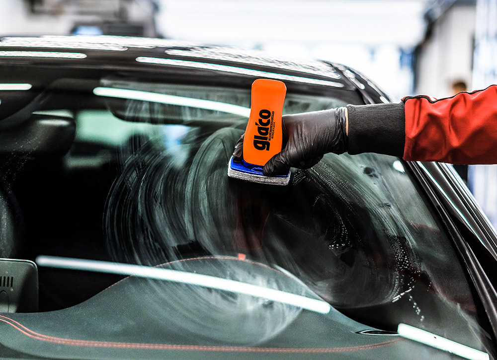 Photo of a car's windscreen being coated with Soft99's Glaco DX glass coating. The product's bottle with integrated felt applicator glides smoothly on the glass surface.